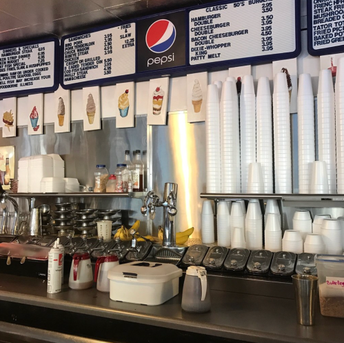 A diner counter with a Pepsi sign, menu items listed, ice cream cones, and stacked cups and containers.