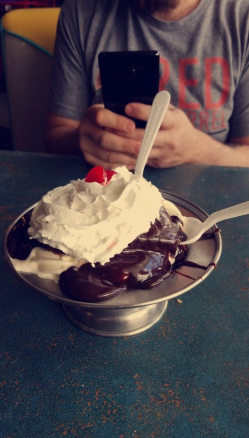 A dessert bowl with chocolate sauce, whipped cream, and a cherry, with a person holding a phone in the background.