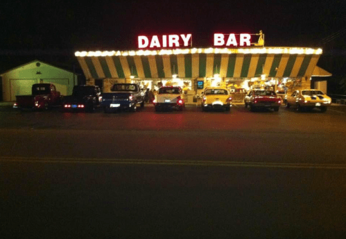 A brightly lit dairy bar at night, featuring vintage cars parked in front under colorful awnings.