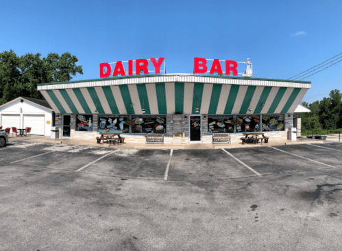 A colorful Dairy Bar with a striped awning, outdoor seating, and a variety of menu items displayed on the windows.