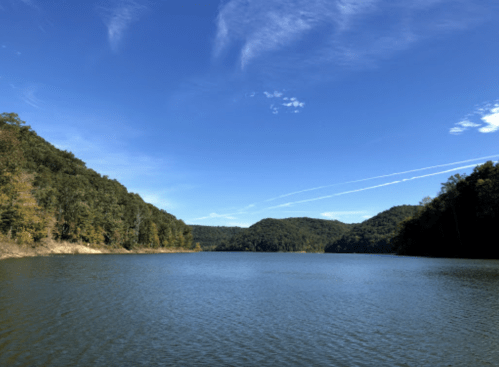 A serene lake surrounded by green hills under a clear blue sky with wispy clouds.