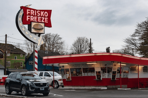 A retro diner called Frisko Freeze with a bright sign, featuring a car parked in front and cloudy skies above.