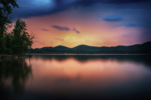 A serene lake at sunset, reflecting colorful skies and surrounded by trees and distant mountains.
