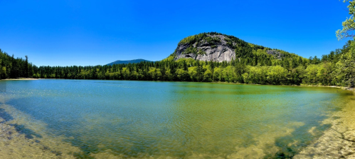 A serene lake surrounded by lush greenery and a rocky hill under a clear blue sky.