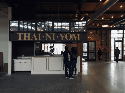 A modern restaurant interior featuring a counter with the sign "THAI·NI·YOM" and patrons standing nearby.
