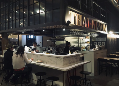 A cozy Thai restaurant with a marble counter, patrons dining, and chefs visible in the open kitchen.