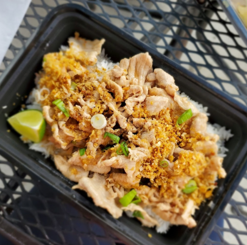 A close-up of a dish featuring sliced meat on rice, topped with crispy garlic, green onions, and a lime wedge.