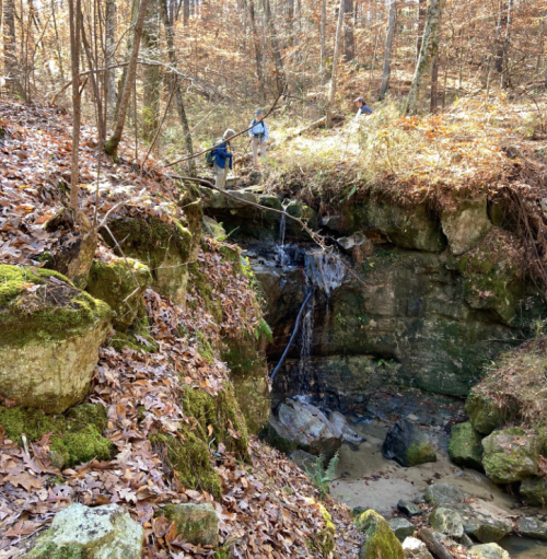 Two people explore a rocky area with a small waterfall, surrounded by autumn leaves and trees in a forest.