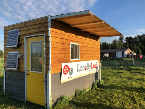 A small wooden building with solar panels, featuring a yellow door and a sign that reads "Locally Laid." Green fields surround it.