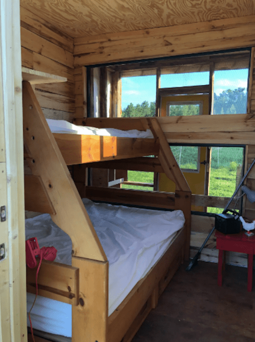 Interior of a wooden cabin with bunk beds, a bright yellow door, and large windows letting in natural light.