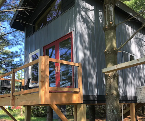A modern treehouse with a red door, elevated on stilts, surrounded by trees and sunlight.