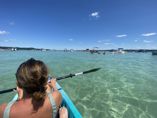 A person kayaking in clear water with boats and people enjoying a sunny day at the beach in the background.