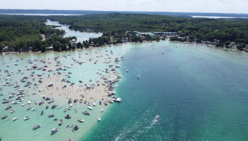 Aerial view of a busy lake with numerous boats and people on the shore, surrounded by lush green trees and hills.