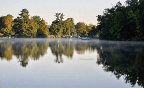 A serene lake at dawn, surrounded by trees, with mist rising from the water and reflections of the landscape.