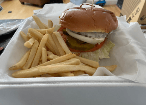 A hamburger with lettuce, tomato, pickles, and onions, served with a side of golden French fries in a takeout container.