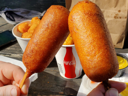 Two golden-brown corn dogs held up, with a side of crispy snacks and a Coca-Cola cup in the background.