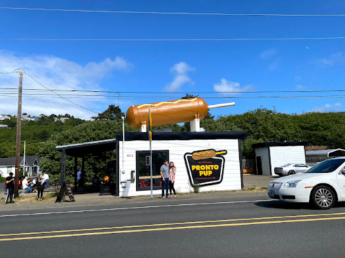 A roadside stand named "Pronto Pup" with a giant corn dog on the roof, people outside, and a car passing by.
