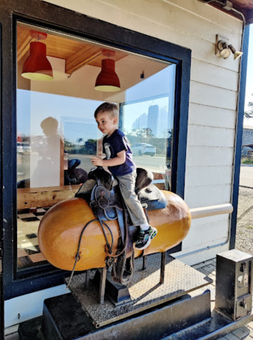 A young boy sits on a decorative saddle outside a building, holding a cup, with a window reflecting the surroundings.