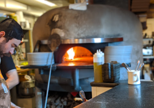 A chef prepares food near a large, traditional wood-fired oven with flames visible inside.