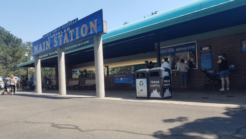 Main station entrance with people waiting in line, benches, and trash bins in a sunny outdoor setting.