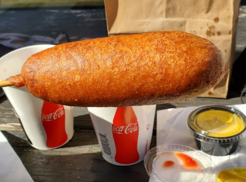 A golden-brown corn dog on a stick, placed between two cups with Coca-Cola branding and dipping sauces.