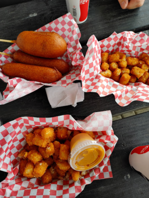 Three baskets of food: two with corn dogs and one with tater tots and cheese sauce, all in red-checkered paper.