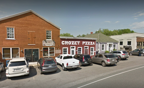 Exterior view of Crozet Pizza and nearby buildings, with parked cars along the street on a sunny day.