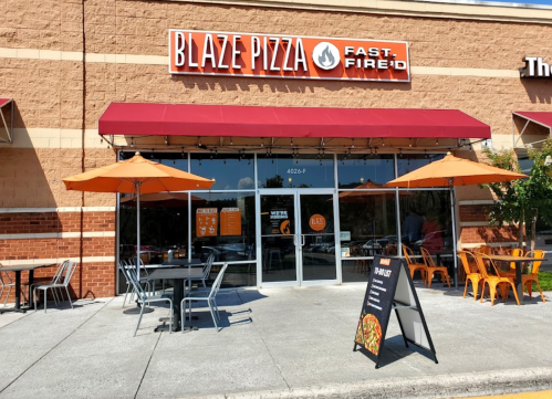 Exterior of Blaze Pizza with orange umbrellas, outdoor seating, and a sign displaying menu options.