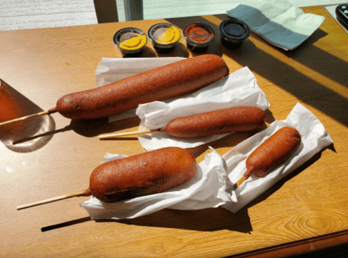 A wooden table with three corn dogs of varying sizes and four dipping sauces in small containers.
