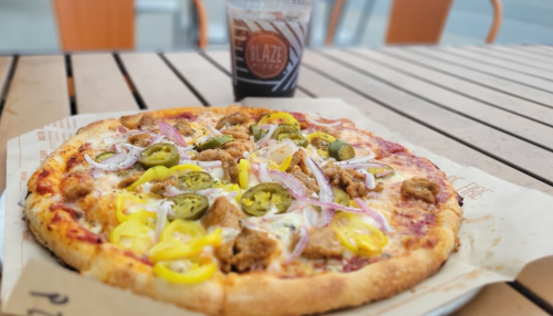 A pizza topped with jalapeños, onions, yellow peppers, and meat, alongside a drink on a wooden table.