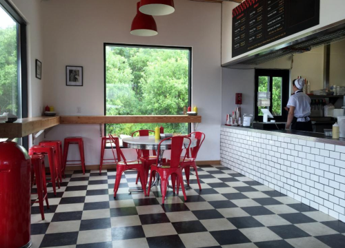 A modern diner with red stools, a checkered floor, and large windows overlooking greenery. A staff member is at the counter.