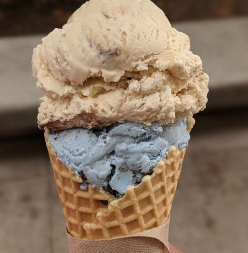 A hand holding a large ice cream cone with two scoops: one light brown and one blue.
