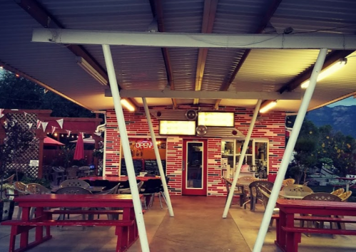 Outdoor dining area with red picnic tables, a colorful brick building, and string lights in the evening.