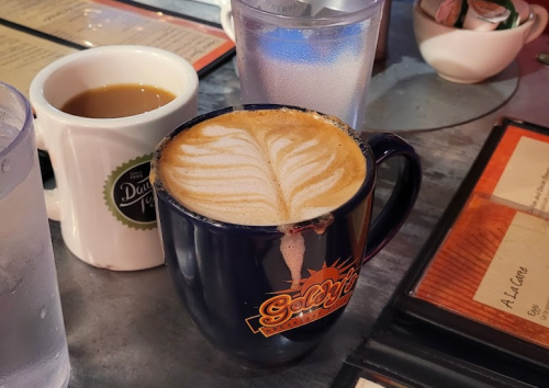 A blue mug with latte art sits on a table next to a white coffee cup and a glass of water.