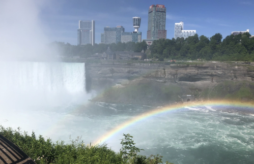 A view of Niagara Falls with a rainbow over the water and city buildings in the background.