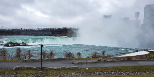 A view of Niagara Falls with mist rising over the water, surrounded by trees and a cloudy sky.