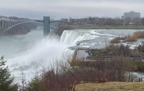 A view of Niagara Falls with rushing water, a bridge in the background, and a cloudy sky above.