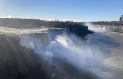 A wide view of a waterfall cascading over cliffs, with mist rising and a clear blue sky above.