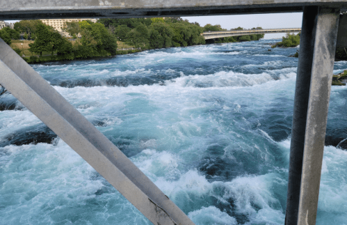 Rushing blue water flows beneath a metal bridge, surrounded by greenery and a distant road.