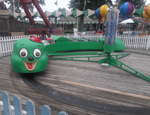 A green cartoonish ride with a smiling face, surrounded by a white picket fence at a carnival.