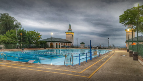 A serene outdoor swimming pool surrounded by trees and buildings, under a cloudy sky at dusk.