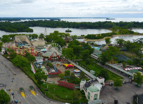 Aerial view of an amusement park with rides, greenery, and a lake in the background under a cloudy sky.