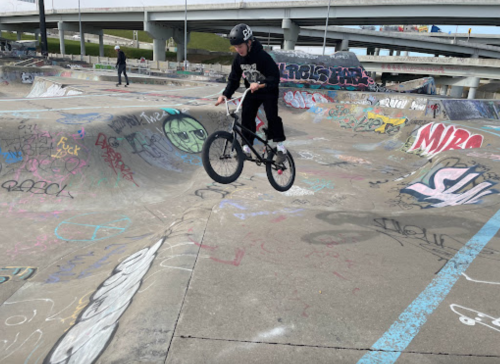 A BMX rider performs a jump at a skatepark filled with graffiti and ramps.