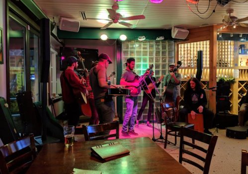 A lively band performs with guitars and percussion in a cozy venue, surrounded by warm lighting and wooden decor.