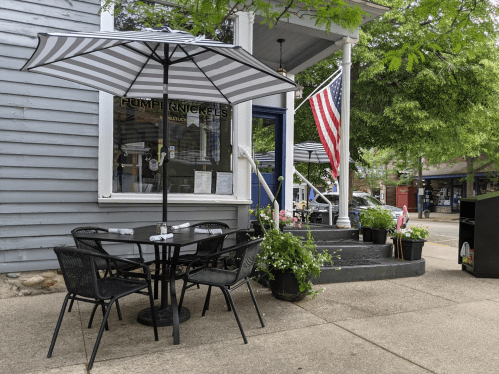 A cozy outdoor café setting with a striped umbrella, black tables, and an American flag near a charming storefront.