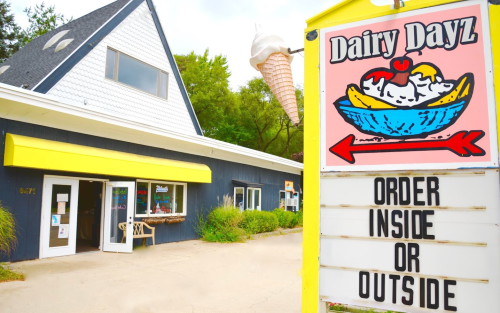 A colorful sign for Dairy Dayz ice cream shop, featuring a sundae graphic, with a building and outdoor seating visible.
