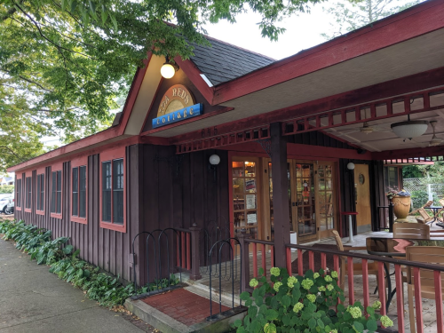 A cozy wooden building with a red trim, featuring large windows and a welcoming entrance surrounded by greenery.