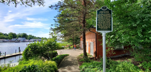 A scenic view of a river with greenery, a historical marker, and a brick building along the water's edge.