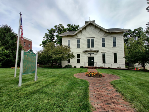 Historic building with a green lawn, flower bed, and a plaque, set against a cloudy sky. An American flag is visible.