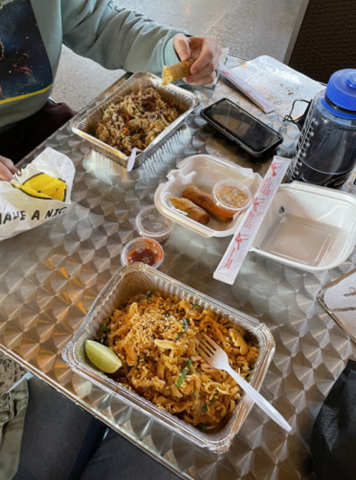 Two takeout containers of food on a table, with a hand holding a spring roll and a drink nearby.